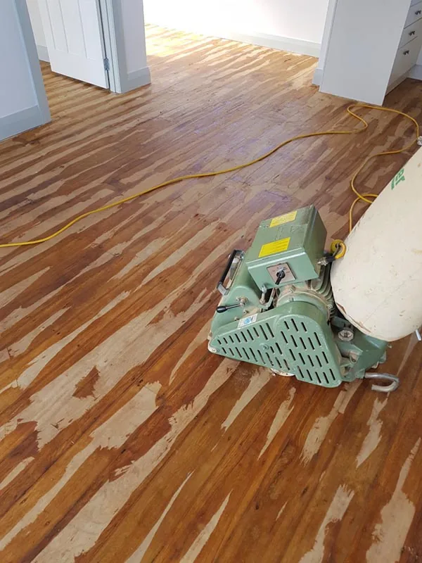 Belt sander on a floor part way through repair, pale epoxy filler visible in the gaps and nail holes between boards
