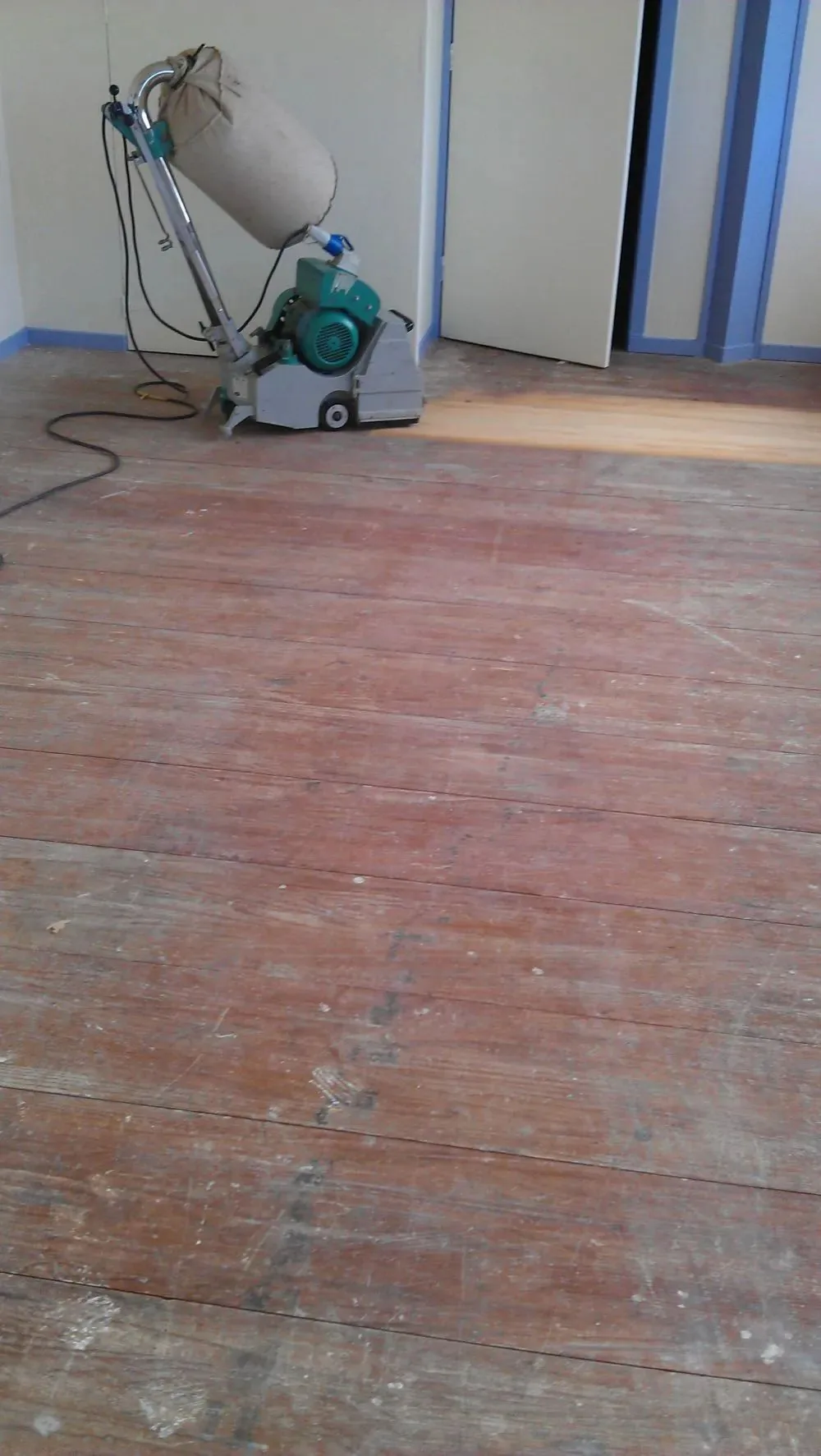 Sanding machine at work on a dark weathered floor, a pale strip of clean timber opening up behind it