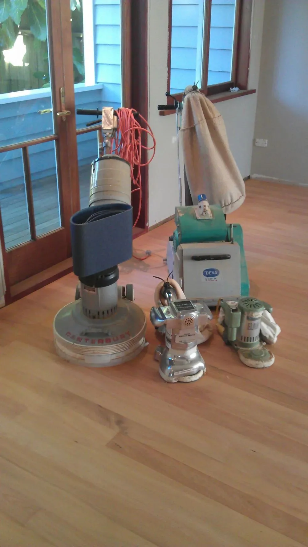 Rotary floor sander and equipment on a warm timber floor beside the blue french doors of an older villa