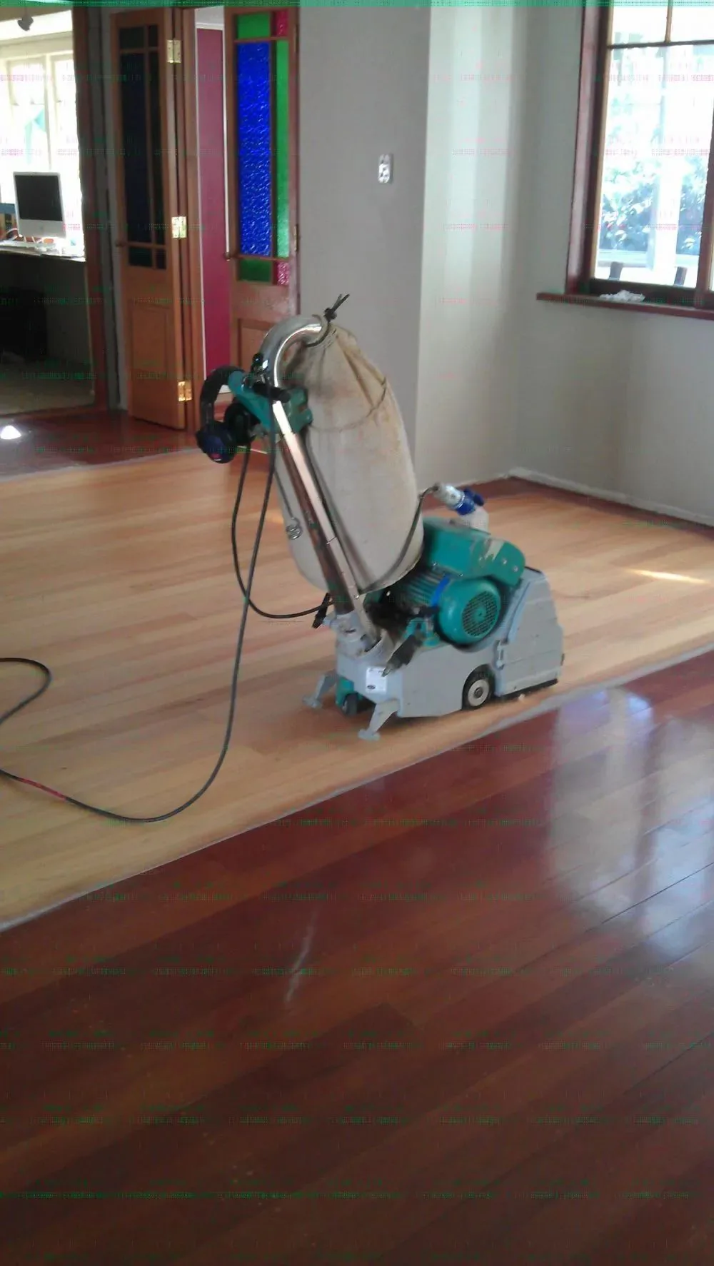 Belt sander part way across a timber floor, half sanded pale and half still dark, with a stained glass door behind